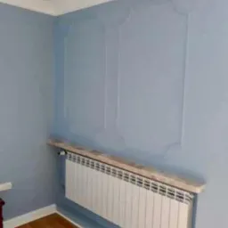 Niebieska ściana z dekoracyjnymi panelami pionowymi, biały grzejnik z drewnianą półką, parkiet na podłodze.