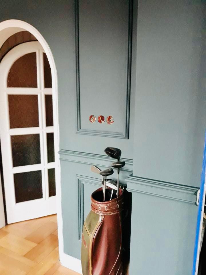 Szaro-niebieska ściana z listwami dekoracyjnymi, trzema puszkami elektrycznymi bez osprzętu i torbą golfową z kijami, widoczna biała, łukowa stolarka drzwiowa w tle.