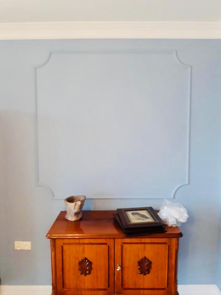 Ściana pomalowana na jasny niebieski kolor z ozdobnym kwadratem z listew, poniżej drewniana komoda z dekoracjami.
