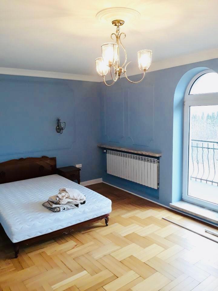 Pokój z niebieskimi ścianami, drewnianą podłogą w jodełkę, łóżkiem z materacem i ozdobnym żyrandolem. Ściany z dekoracyjnymi listwami i grzejnikiem pod parapetem.