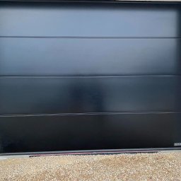 AQ-ELTECH - Zamknięta, czarna, segmentowa, automatyczna brama z poziomymi przetłoczeniami, widoczne logo producenta w prawym dolnym rogu, podłoże wysypane drobnym żwirem.