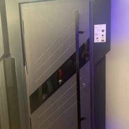 AQ-ELTECH - Nowoczesne, szare drzwi wejściowe z długim, pionowym uchwytem i dekoracyjnymi, ukośnymi liniami, prezentowane w salonie wystawowym pod logiem producenta 'MARTOM'.