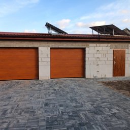 AQ-ELTECH - Garaż z dwoma brązowymi bramami segmentowymi i brązowymi drzwiami wejściowymi, budynek w trakcie budowy z białego bloczka, panele słoneczne na dachu, szara kostka brukowa na podjeździe.