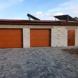 Garaż z dwoma brązowymi bramami segmentowymi i brązowymi drzwiami wejściowymi, budynek w trakcie budowy z białego bloczka, panele słoneczne na dachu, szara kostka brukowa na podjeździe.