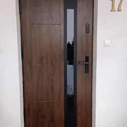 Brązowe drzwi wejściowe z czarnym pionowym przeszkleniem, numer domu 17 na białej elewacji, widoczny fragment chodnika z cegły