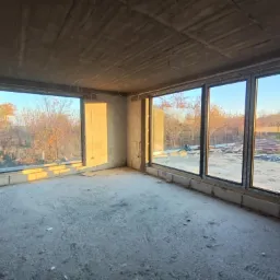 Surowy stan wnętrza z trzema dużymi oknami z czarnymi ramami, widok na jesienne drzewa i plac budowy na zewnątrz, surowy betonowy sufit, ściany z pustaków, wpadające promienie słoneczne.