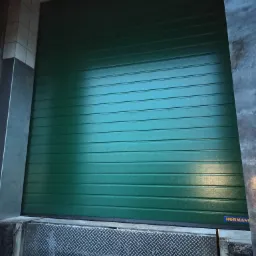 Zamknięta, zielona brama segmentowa z poziomymi przetłoczeniami, widoczna z bliska, z fragmentem rampy załadunkowej i logo producenta w prawym dolnym rogu.