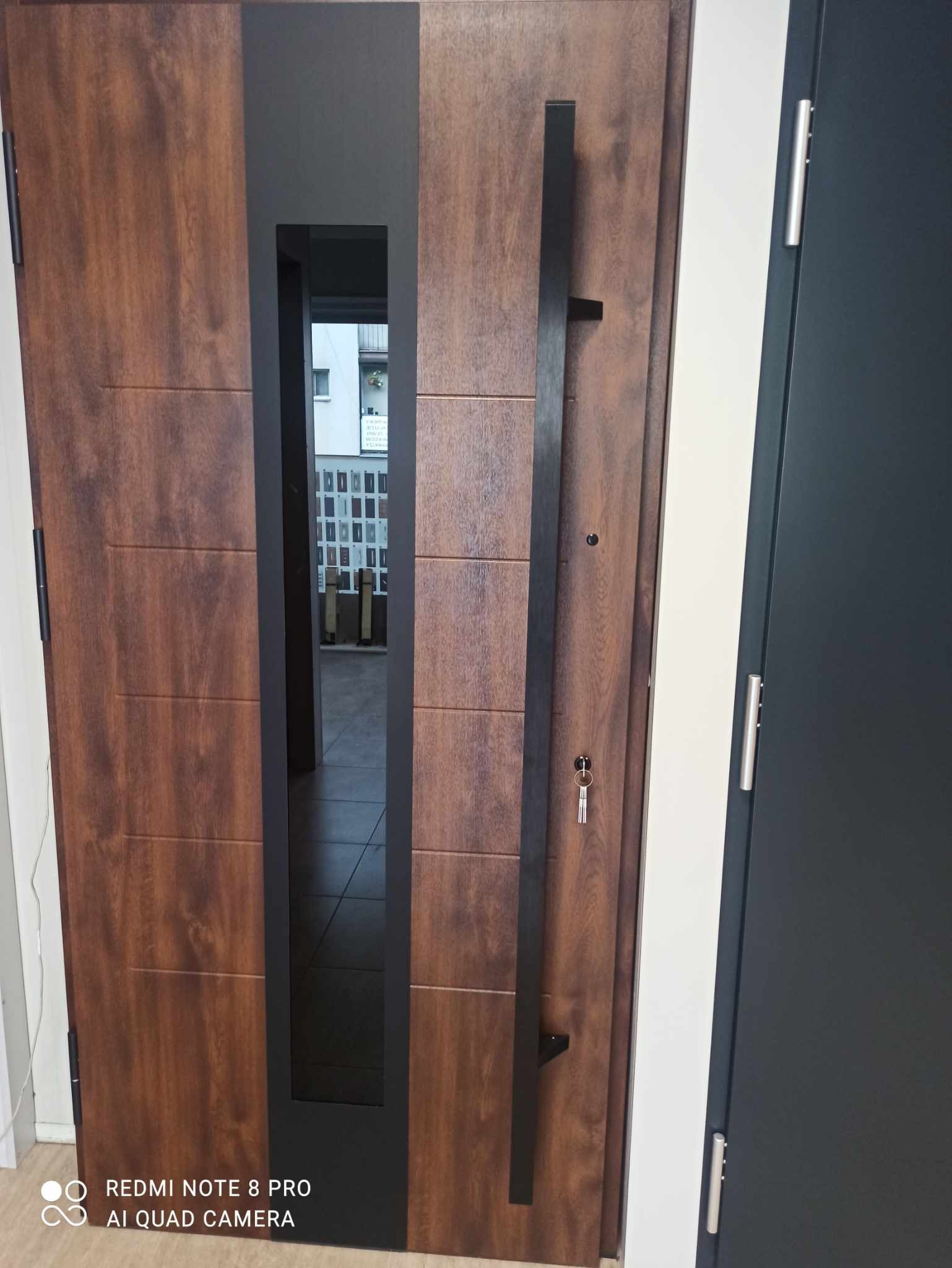 Nowoczesne drzwi wejściowe w odcieniach brązu z czarnym, pionowym uchwytem i przeszkleniem, widok z bliska na klucz w zamku.