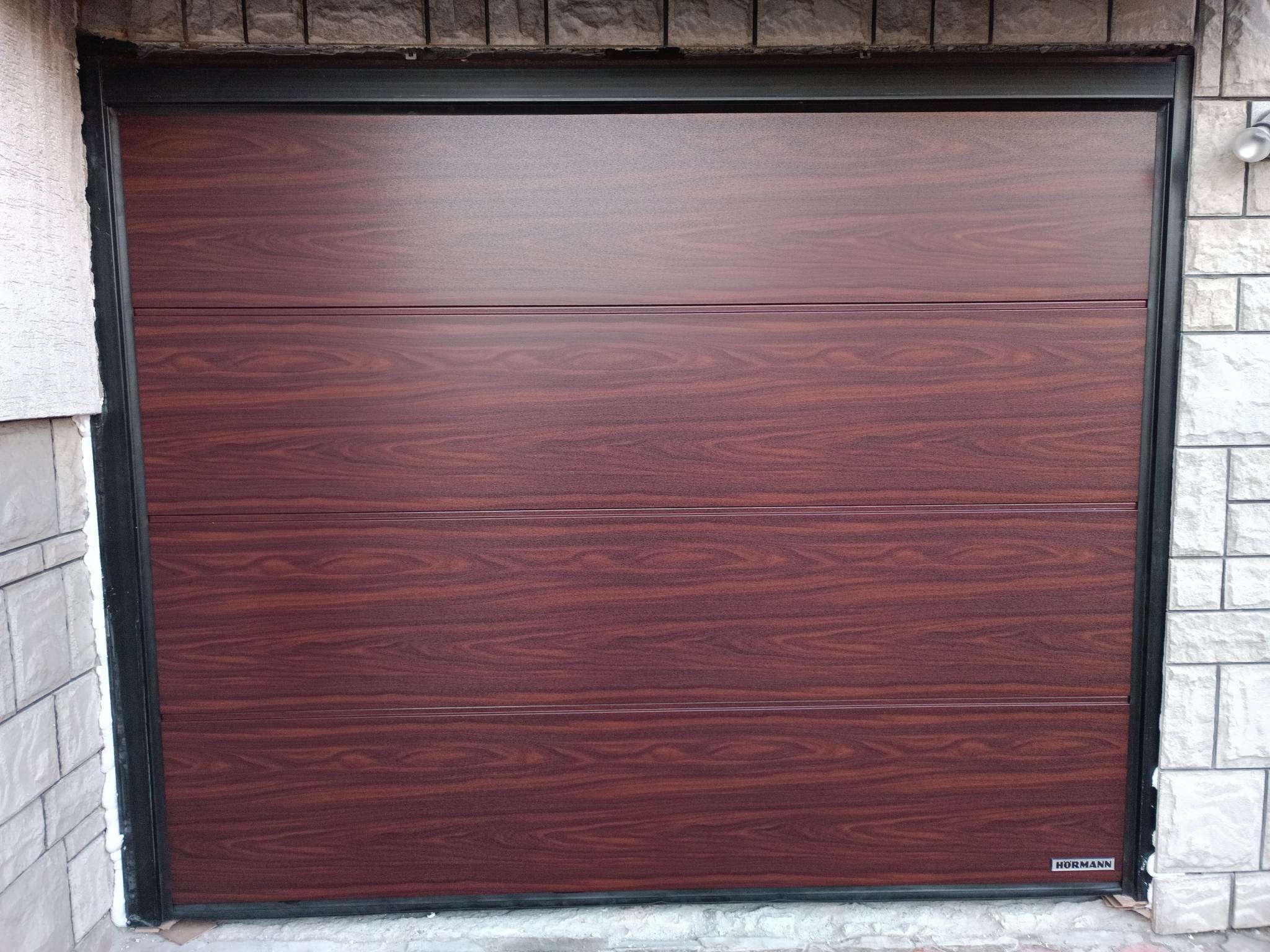 Zamknięta, segmentowa brama garażowa w kolorze ciemnego drewna z czarną ramą, zamontowana w elewacji wykończonej jasnym kamieniem dekoracyjnym. Widoczny fragment lampy zewnętrznej.
