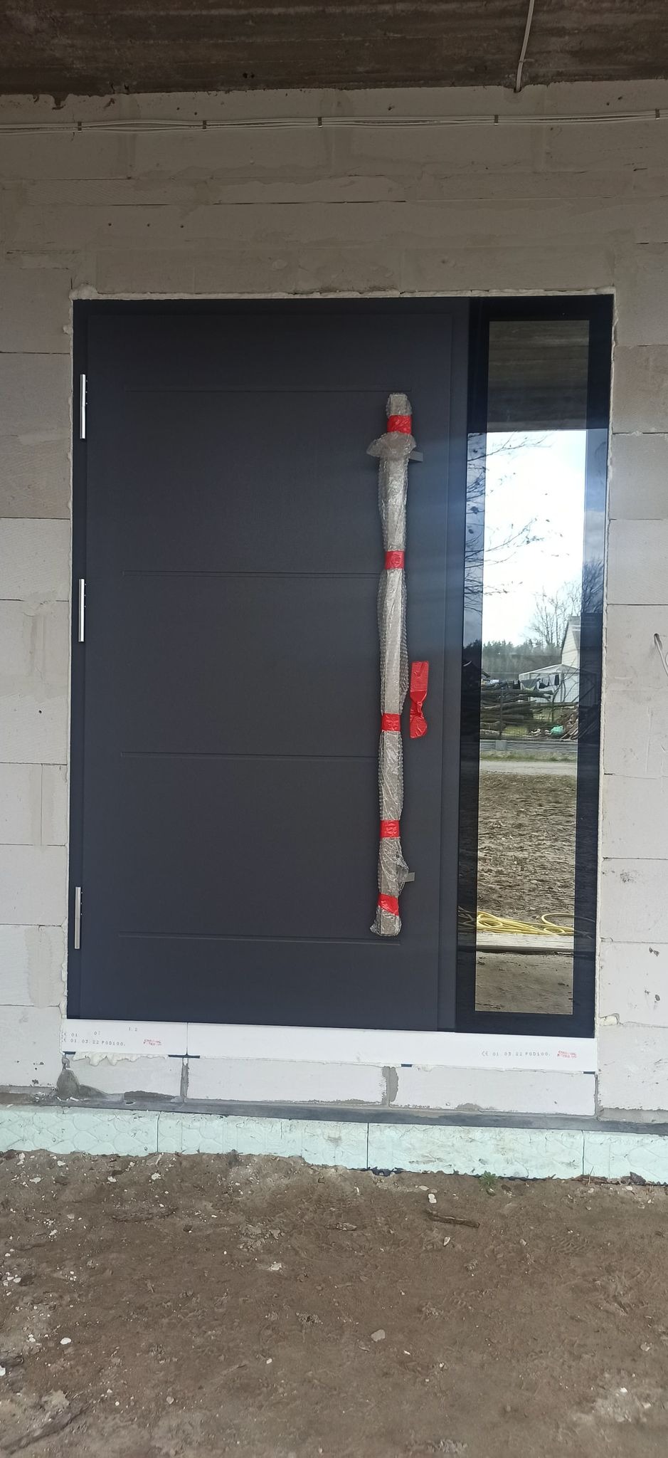 Nowe, szare drzwi wejściowe z bocznym panelem szklanym, jeszcze zabezpieczone folią ochronną na klamce, zamontowane w surowym stanie budynku z bloczków.