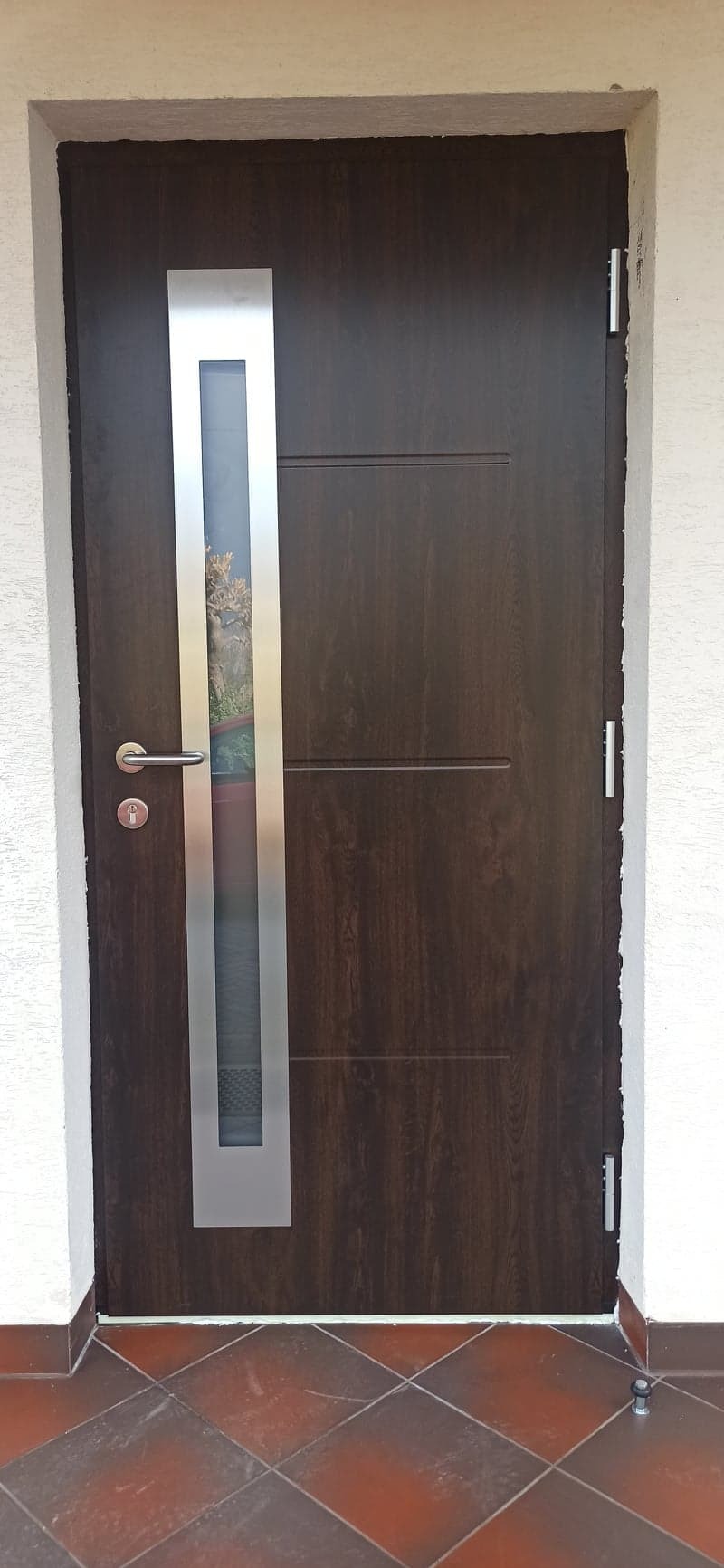 Ciemnobrązowe drzwi wejściowe z pionowym przeszkleniem w srebrnej ramie, widoczny zamek i klamka, na podłodze brązowe płytki.