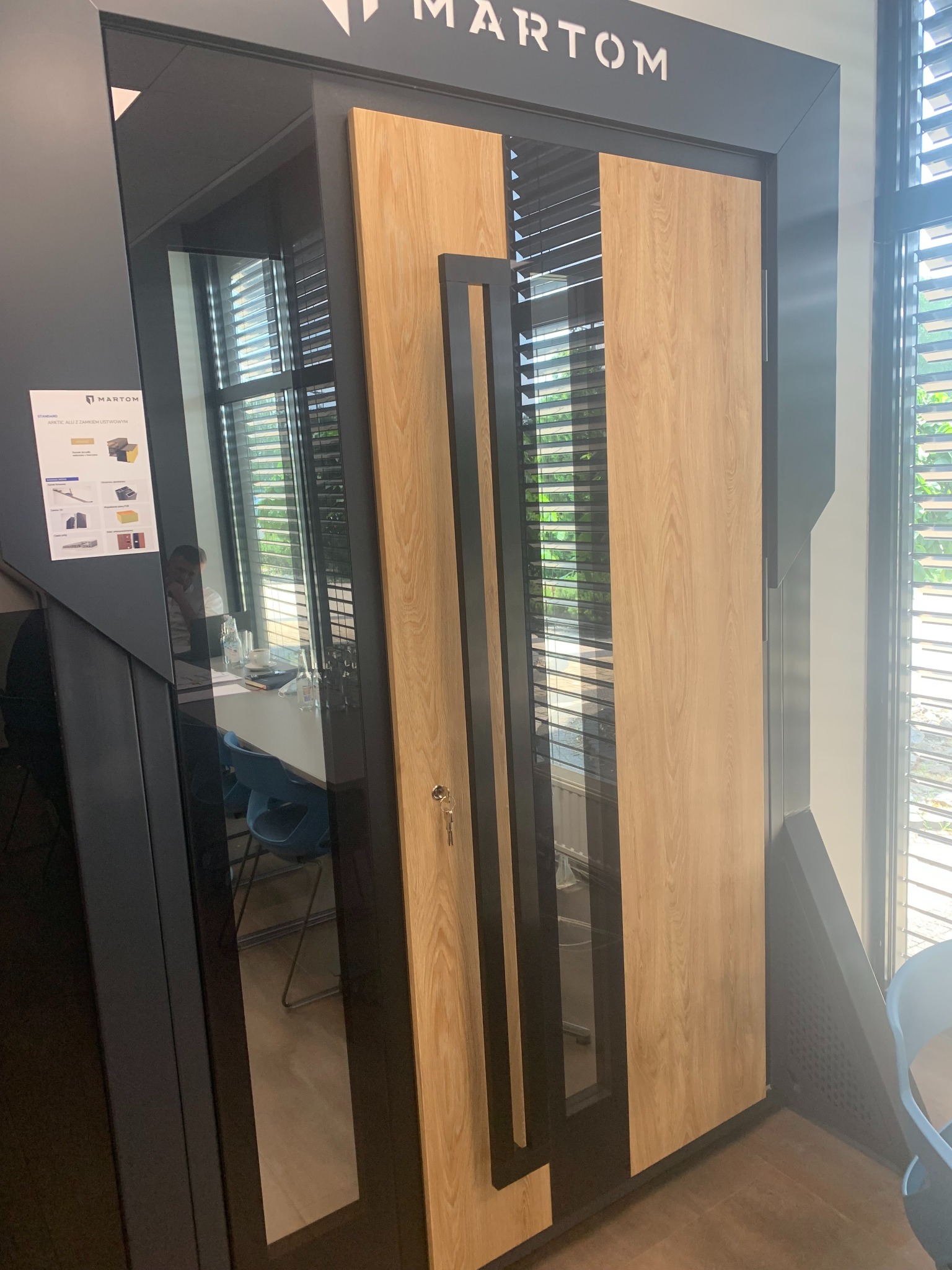 Wystawowa, drewnopodobna, nowoczesna stolarka drzwiowa z czarnym, geometrycznym uchwytem i widocznym kluczem w zamku, na tle biura z żaluzjami.