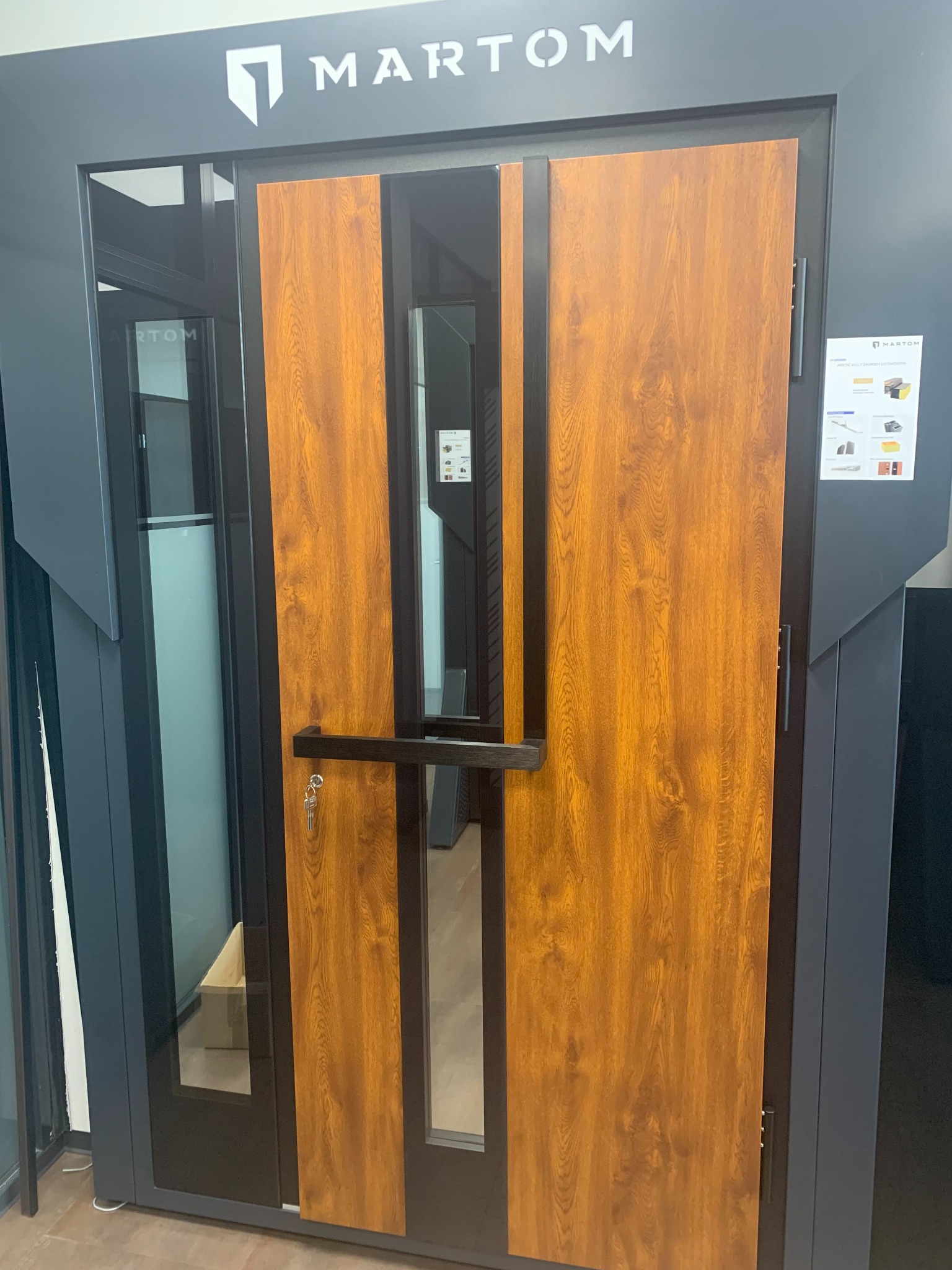 Nowoczesne drzwi zewnętrzne Martom z drewnopodobnym panelem i czarną ramą, prezentowane w salonie sprzedaży z widocznymi kluczami w zamku.