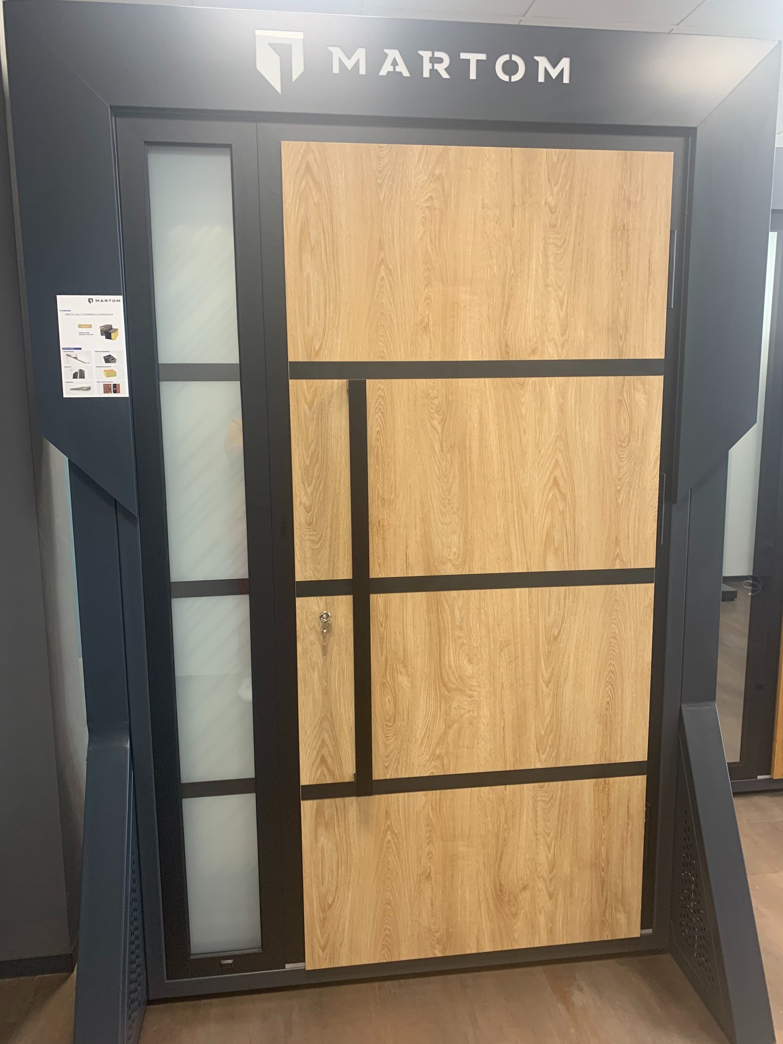 Nowoczesne drzwi zewnętrzne z drewnopodobnym panelem i matowym szkłem, czarna rama, widoczny klucz w zamku i logo producenta Martom.