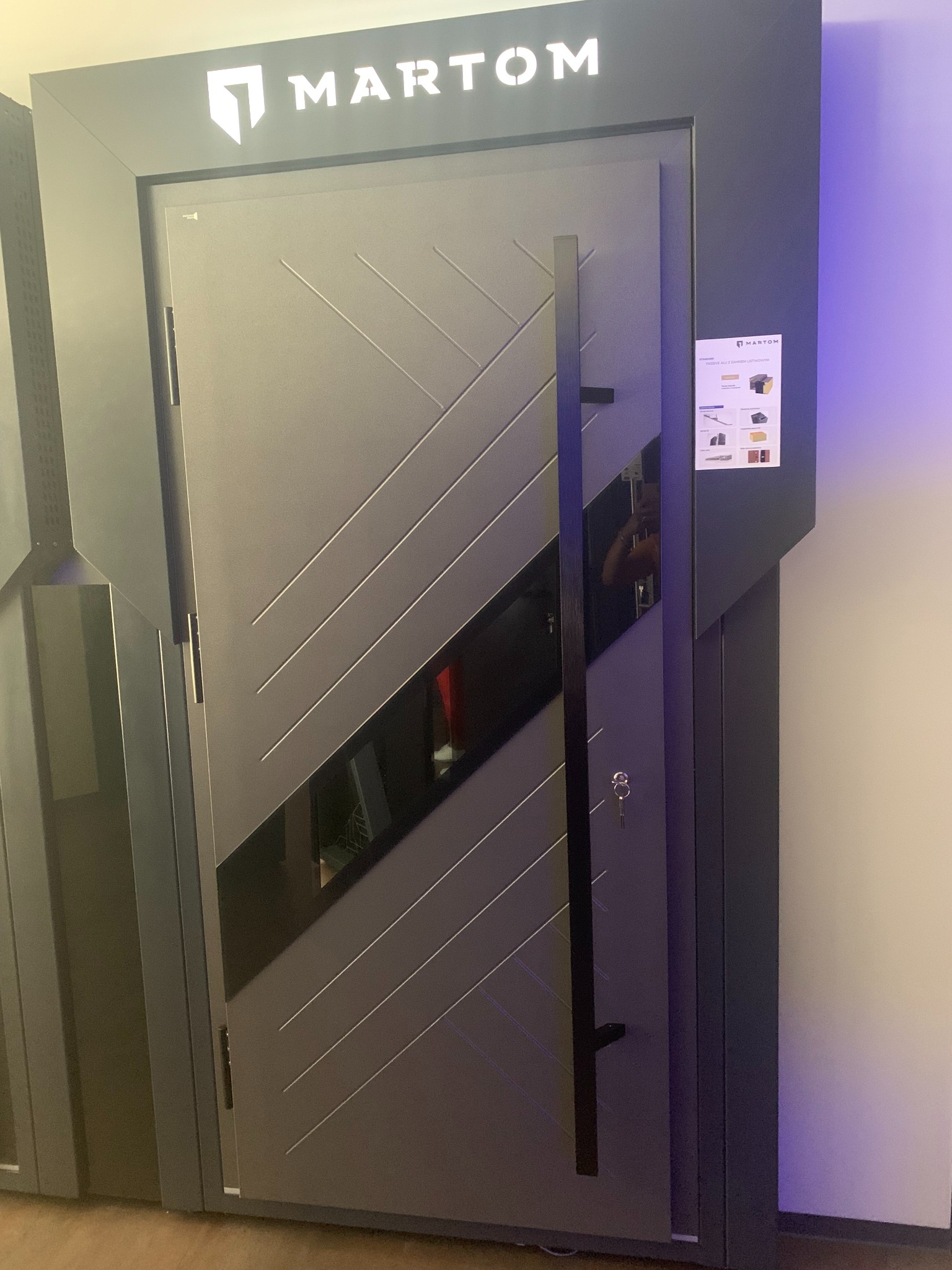 Nowoczesne, szare drzwi wejściowe z długim, pionowym uchwytem i dekoracyjnymi, ukośnymi liniami, prezentowane w salonie wystawowym pod logiem producenta 'MARTOM'.