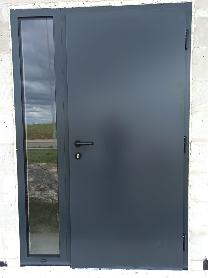 Grafitowe drzwi wejściowe z bocznym przeszkleniem, zamontowane w surowym murze z betonu komórkowego.