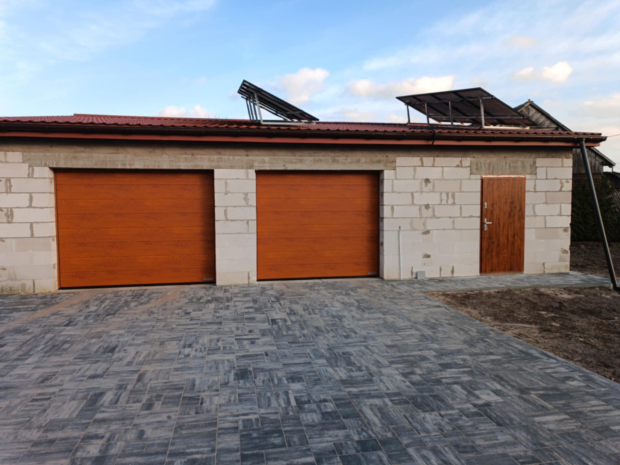 Garaż z dwoma brązowymi bramami segmentowymi i brązowymi drzwiami wejściowymi, budynek w trakcie budowy z białego bloczka, panele słoneczne na dachu, szara kostka brukowa na podjeździe.