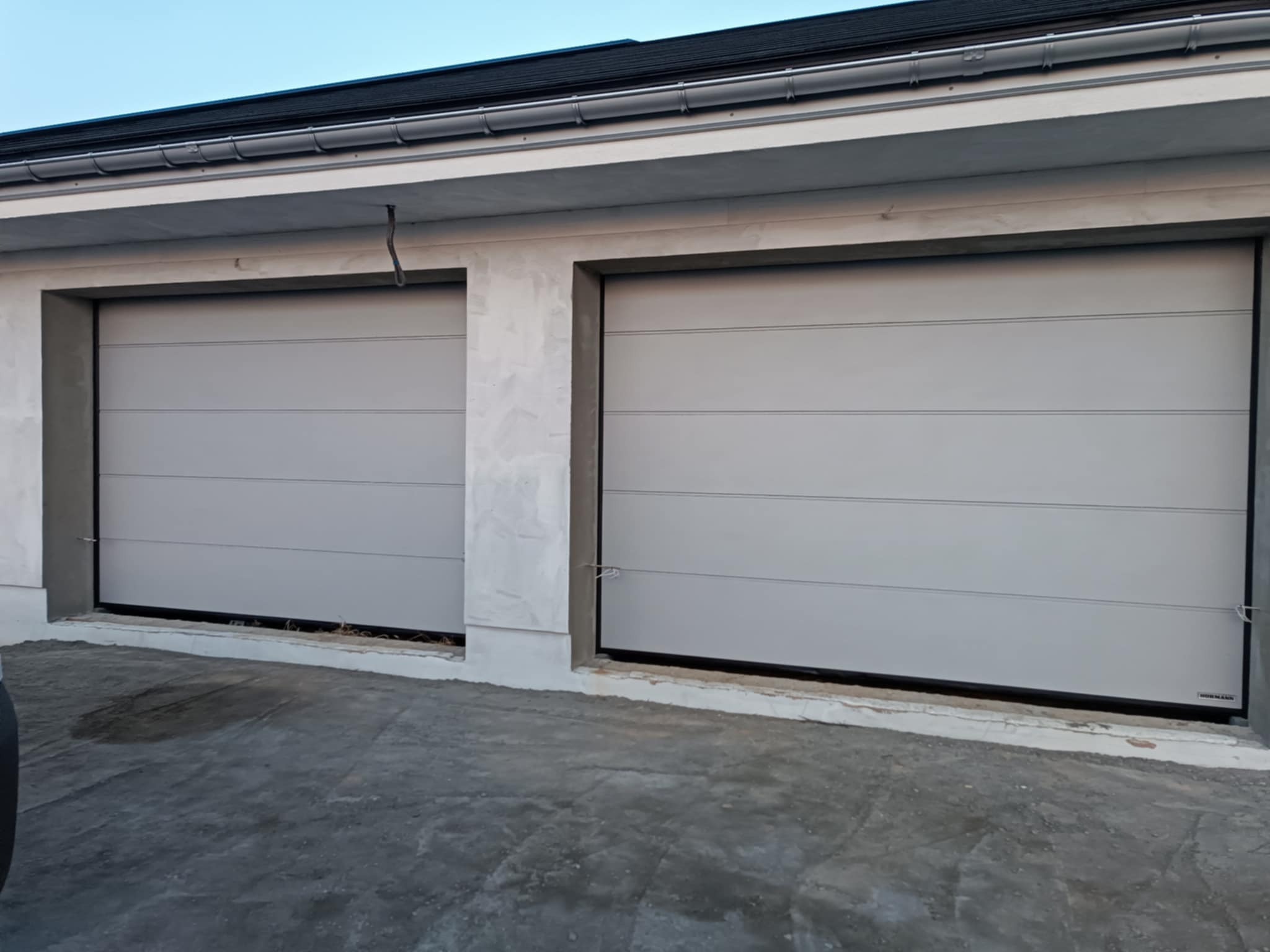 Dwie szare segmentowe bramy garażowe z poziomymi przetłoczeniami, zamontowane w surowym, częściowo otynkowanym garażu z czarnym dachem. Widoczny fragment betonowego podjazdu.