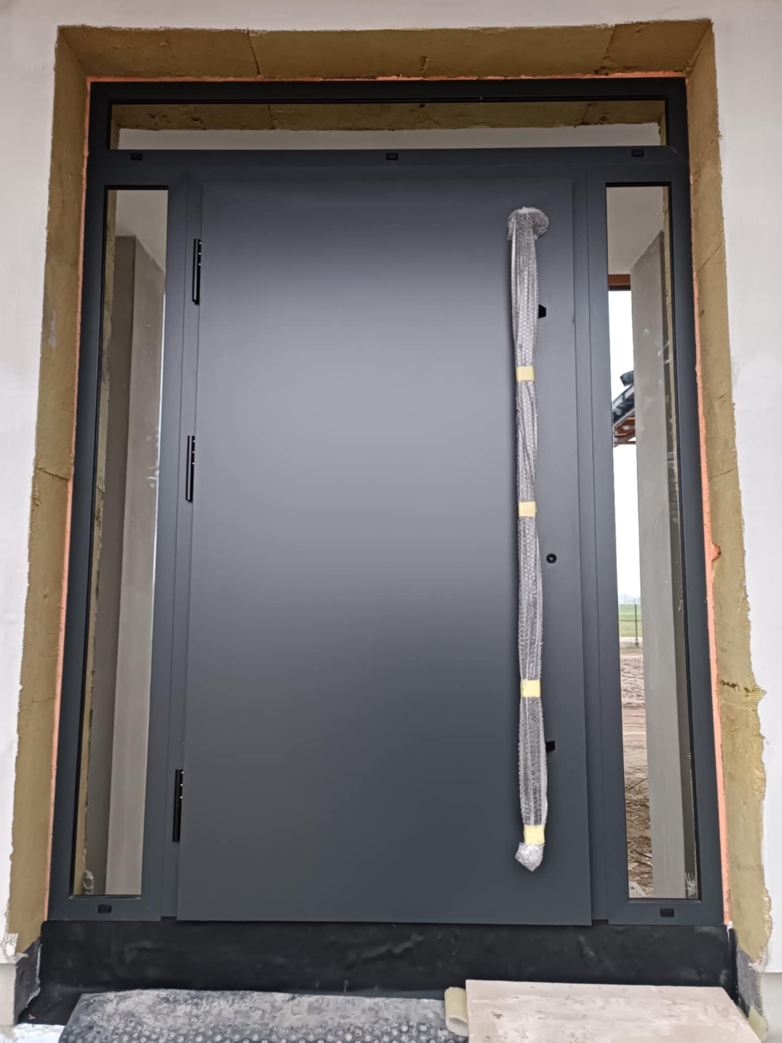 Montaż szarych drzwi wejściowych z bocznymi przeszkleniami, widoczne uszczelnienia i folia ochronna na klamce, ościeżnica osadzona w surowym murze.