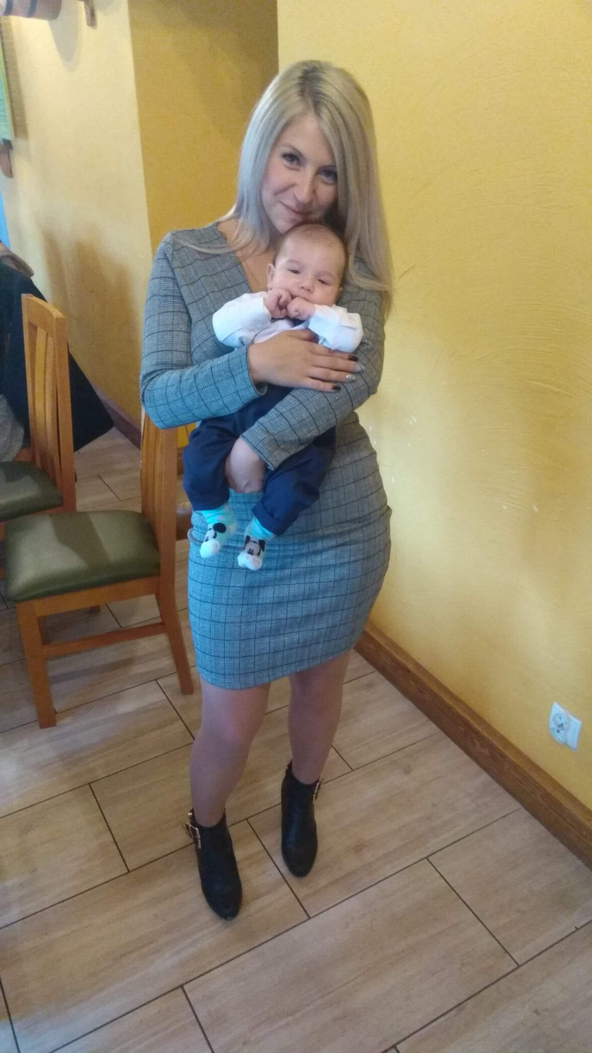 Kobieta w szarej sukience w kratę trzyma na rękach niemowlę w białej koszuli i granatowych spodniach, dziecko ma na sobie skarpetki z motywem Myszki Miki.