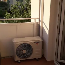 Biała jednostka zewnętrzna klimatyzacji Kaisai na balkonie z brązowymi płytkami, w tle budynek mieszkalny.