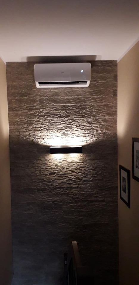 Biała jednostka wewnętrzna klimatyzatora zamontowana na ścianie wyłożonej dekoracyjnym kamieniem, oświetlona od dołu przez minimalistyczną lampę ścienną.