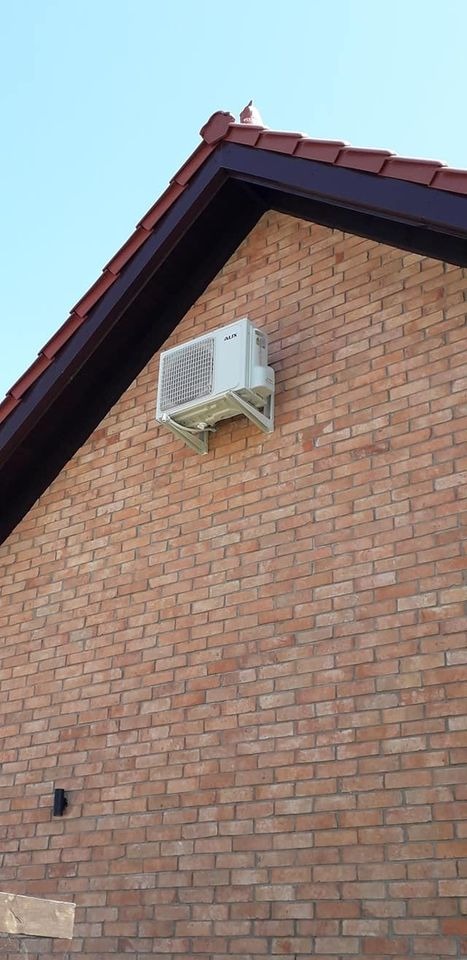 Biała jednostka zewnętrzna klimatyzatora marki AUX zamontowana na wspornikach na ceglanej ścianie budynku z ciemnobrązowym dachem z czerwonej dachówki, widok z dołu.