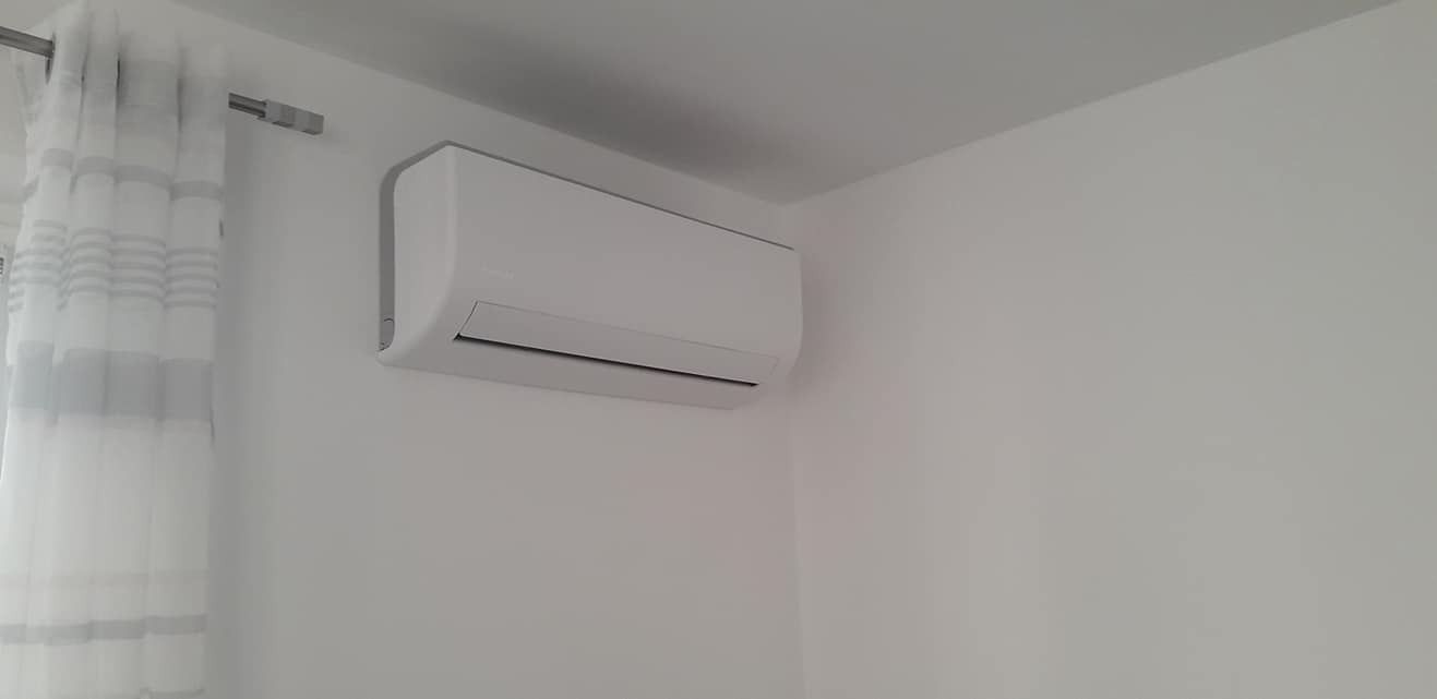 Nowoczesny, biały klimatyzator ścienny zamontowany w jasnym pomieszczeniu z minimalistycznym wystrojem, widoczny fragment białej zasłony w lewym górnym rogu.