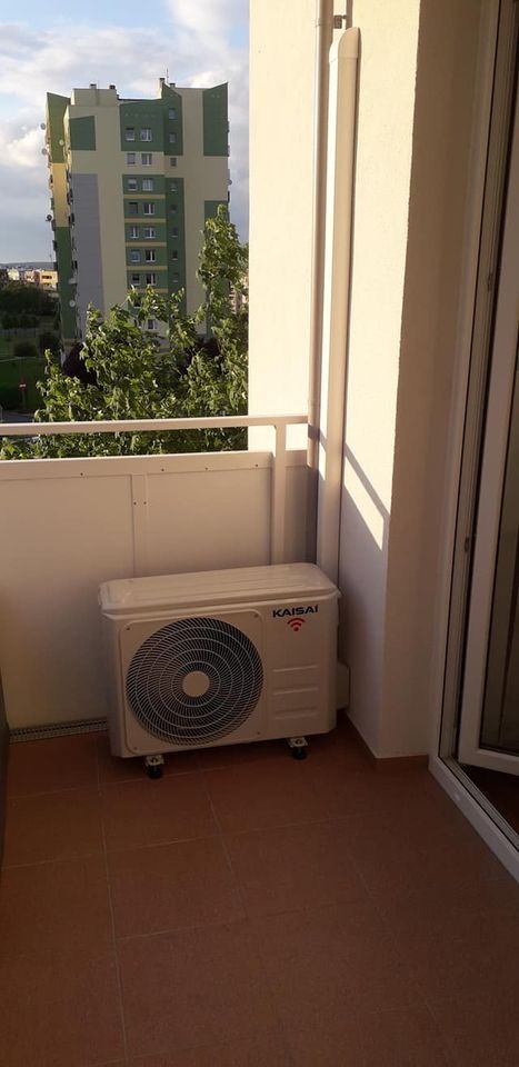 Biała jednostka zewnętrzna klimatyzacji Kaisai na balkonie z brązowymi płytkami, w tle budynek mieszkalny.