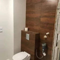 Podwieszana toaleta ze spłuczką ukrytą w obudowie z płytek imitujących drewno, na ścianie rustykalne płytki z geometrycznym wzorem, widoczne przyłącza wodne i kanalizacyjne w trakcie montażu.