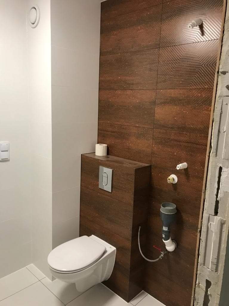 Podwieszana toaleta ze spłuczką ukrytą w obudowie z płytek imitujących drewno, na ścianie rustykalne płytki z geometrycznym wzorem, widoczne przyłącza wodne i kanalizacyjne w trakcie montażu.