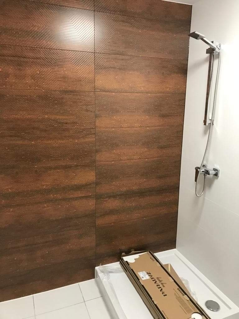 Nowoczesna kabina prysznicowa z brązowymi płytkami imitującymi drewno i chromowaną armaturą, w tle karton z nazwą producenta płytek.