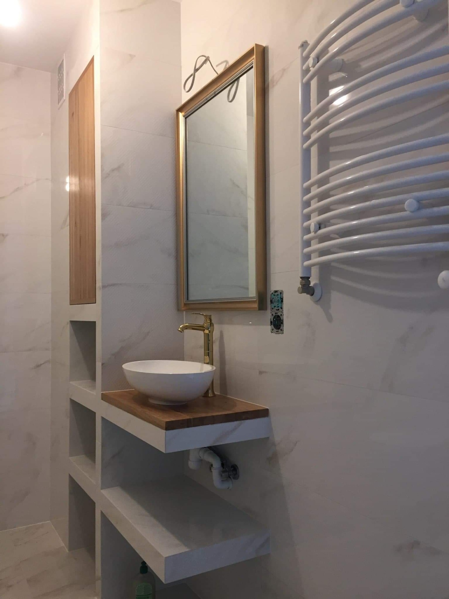 Nowoczesna łazienka z umywalką nablatową na drewnianym blacie, złotą baterią i grzejnikiem drabinkowym na tle marmurowych płytek.