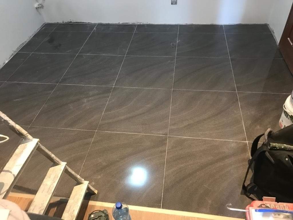 Ułożone ciemnoszare płytki podłogowe z jasnymi fugami, widoczne narzędzia pracy i drabina w trakcie realizacji.