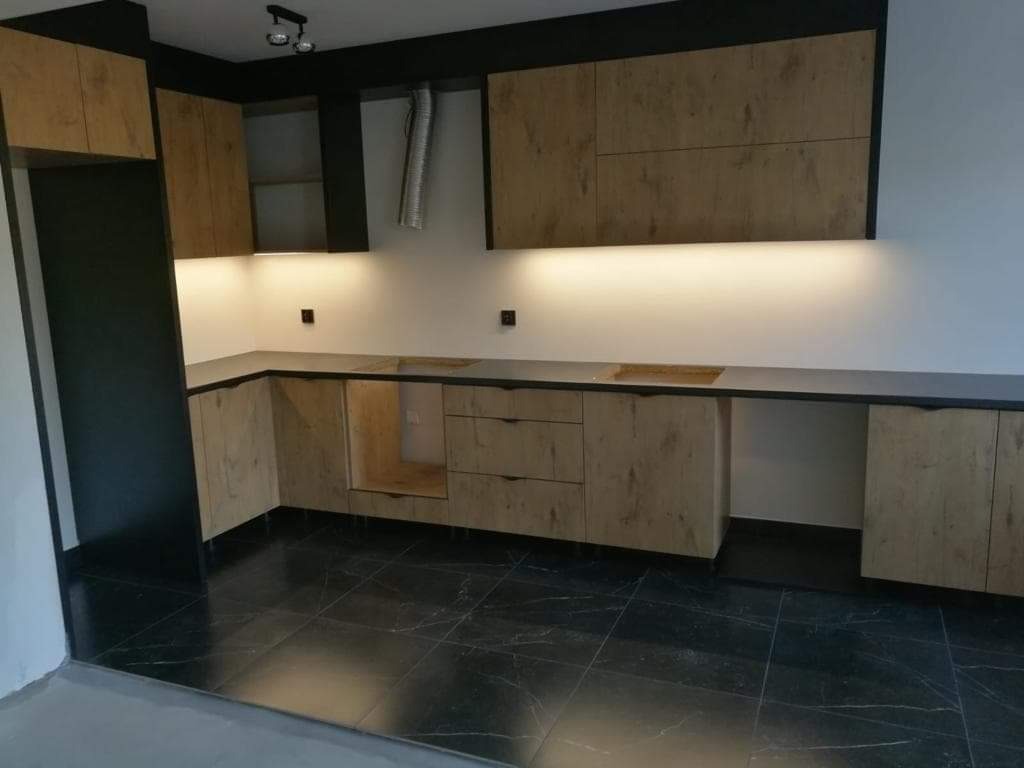 Częściowo zamontowana kuchnia z drewnopodobnymi frontami i czarnymi akcentami, widoczne podświetlenie szafek górnych i czarna podłoga z połyskiem.