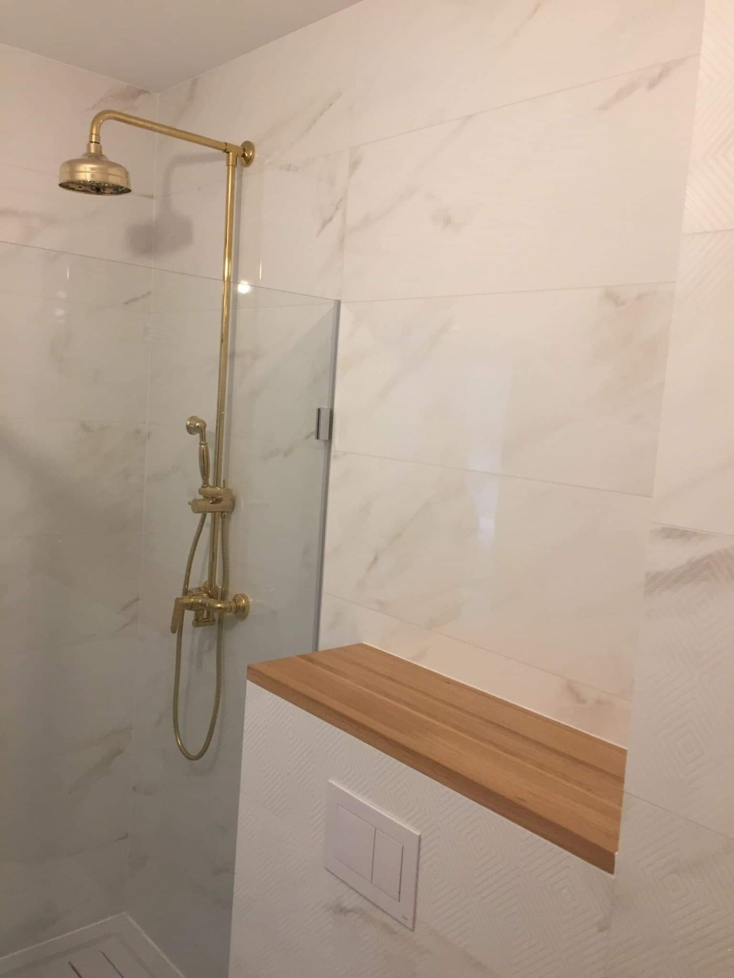 Nowoczesna kabina prysznicowa z deszczownicą i baterią w kolorze złotym, wykończona marmurowymi płytkami i drewnianą półką z ukrytym panelem sterowania.