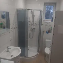 Wnętrze łazienki z kabiną prysznicową, umywalką z szafką, toaletą i oknem. Widoczne białe płytki na ścianach z delikatnym wzorem oraz drewnopodobne płytki na podłodze.