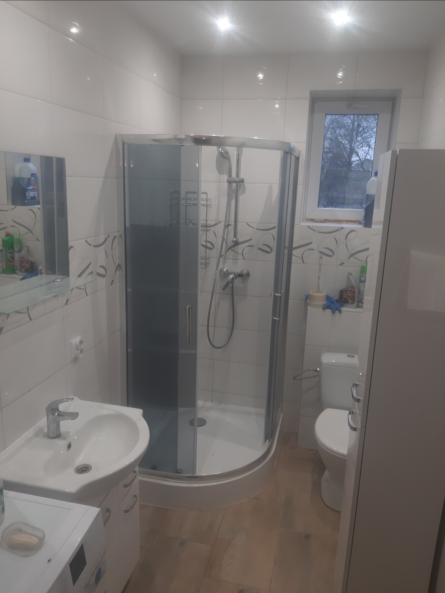 Wnętrze łazienki z kabiną prysznicową, umywalką z szafką, toaletą i oknem. Widoczne białe płytki na ścianach z delikatnym wzorem oraz drewnopodobne płytki na podłodze.