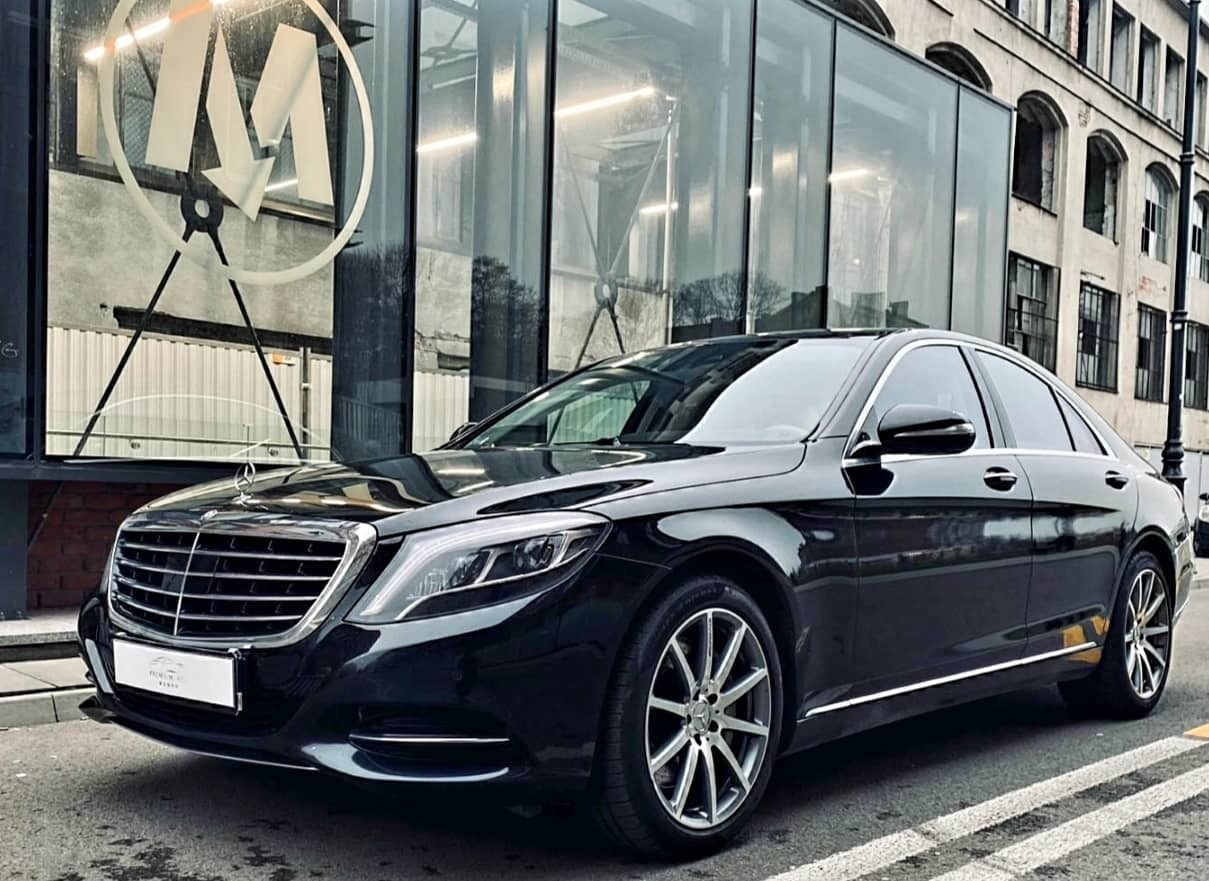 Czarna limuzyna Mercedes S-Class zaparkowana przed nowoczesnym budynkiem z przeszkloną fasadą i dużym logo na szybie.