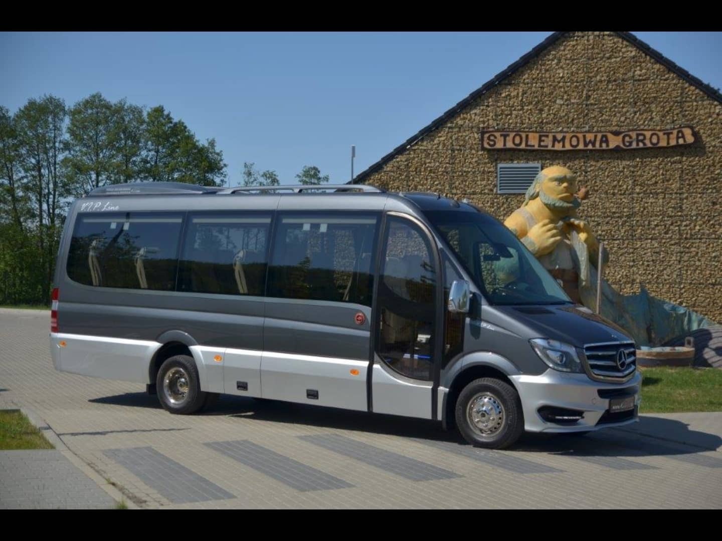 Szaro-srebrny bus Mercedes-Benz z otwartymi drzwiami na tle budynku 'Stolemowa Grota' z charakterystyczną figurą brodatego mężczyzny z fajką.