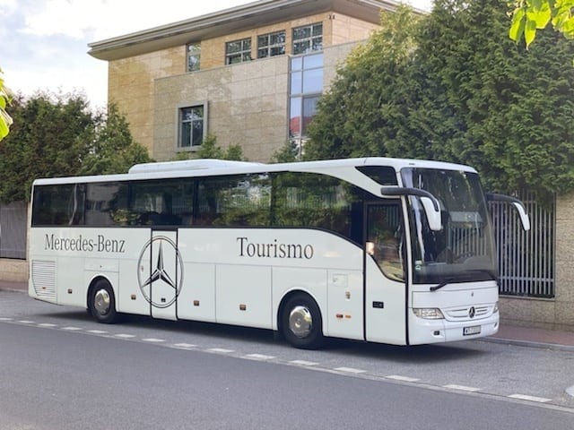 Biały autokar Mercedes-Benz Tourismo zaparkowany na ulicy przed budynkiem z zielonymi drzewami w tle.