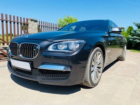 Czarny samochód osobowy BMW serii 7 na podjeździe z kostki brukowej, widoczne ogrodzenie posesji i zieleń w tle.