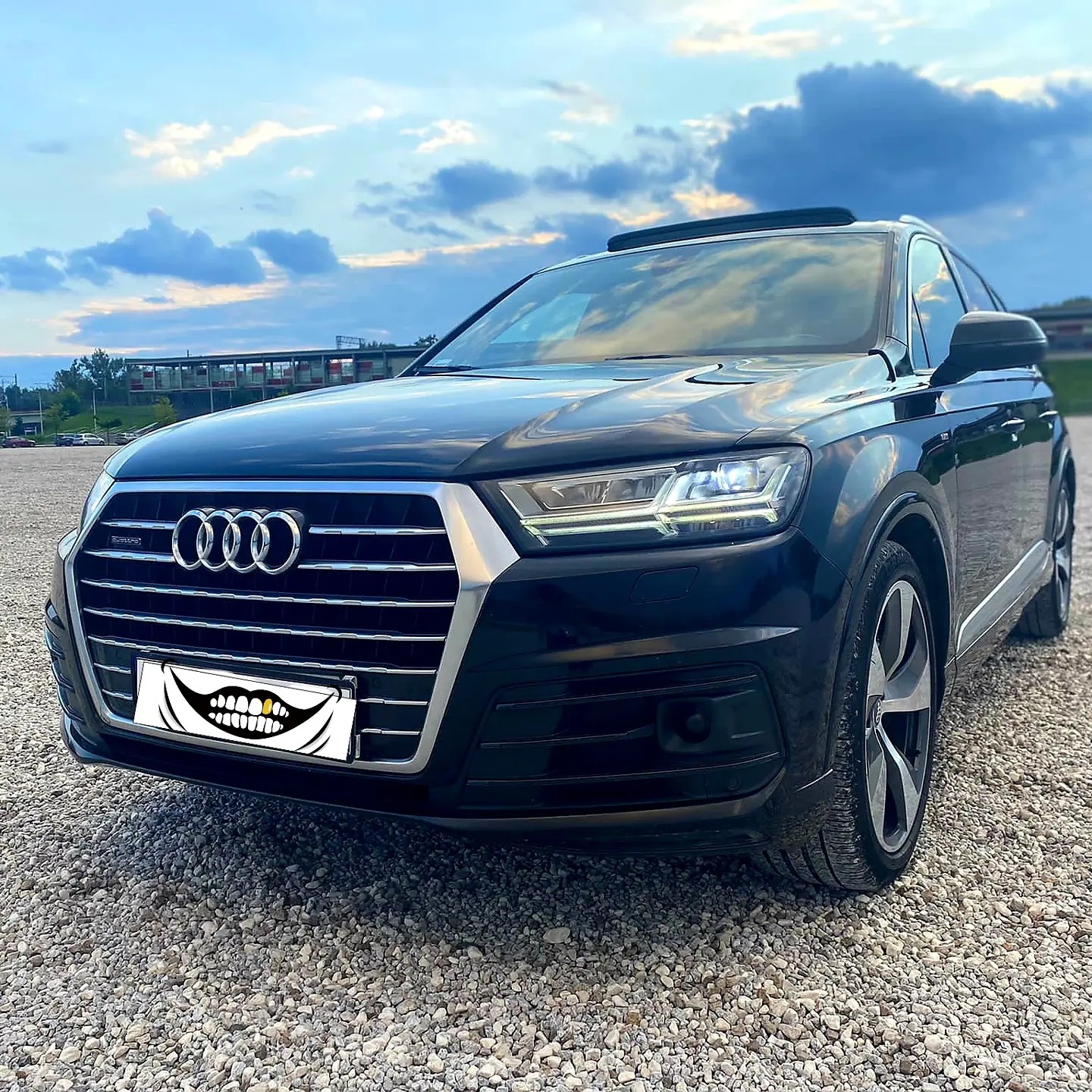 Czarne Audi Q7 z panoramicznym dachem, zaparkowane na żwirowym parkingu, z widocznym budynkiem w tle pod błękitnym, pochmurnym niebem. Na tablicy rejestracyjnej naklejka z uśmiechem.