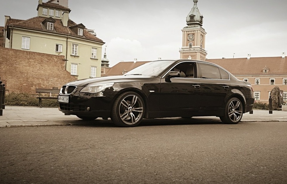 Czarna limuzyna BMW serii 5 zaparkowana na tle Starego Miasta w Warszawie, widoczna Wieża Zegarowa Zamku Królewskiego.