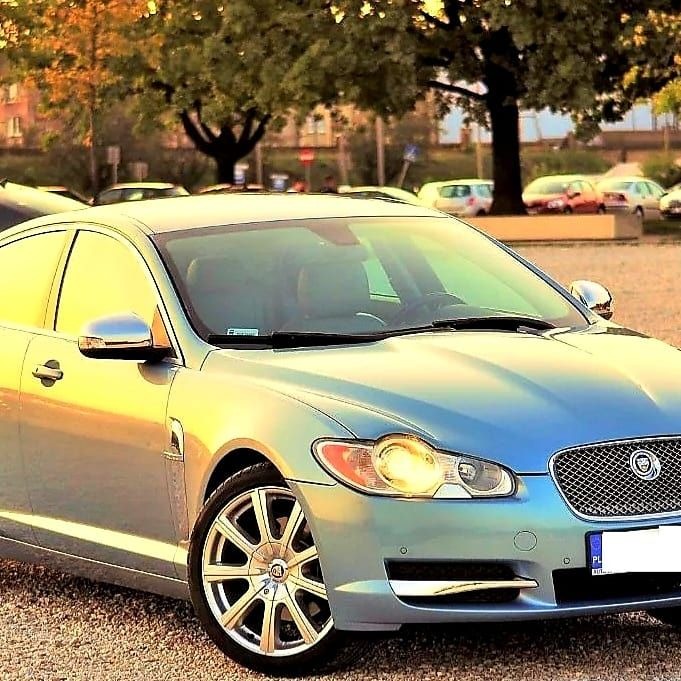 Błękitny Jaguar XF zaparkowany na żwirowym parkingu, z widocznymi budynkami i drzewami w tle, oświetlony ciepłym światłem zachodzącego słońca.