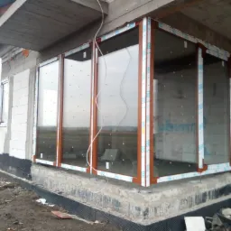 Montaż dużych okien panoramicznych z brązowymi ramami w surowym stanie budynku, widoczne zabezpieczenia na ramach i otaczający plac budowy.