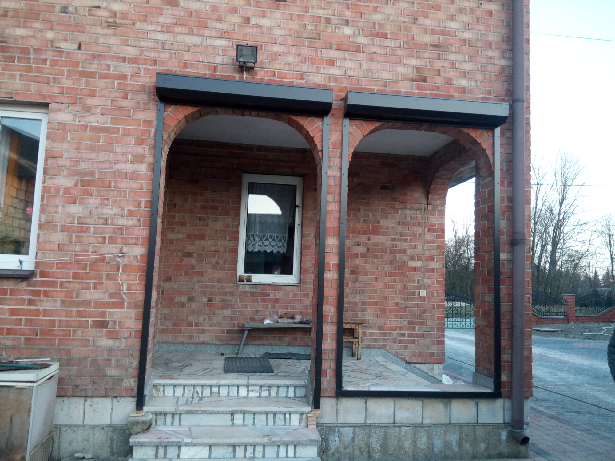 Ceglana fasada budynku z dwoma łukowymi wejściami, gdzie zamontowano czarne rolety zewnętrzne, częściowo zasłonięte.