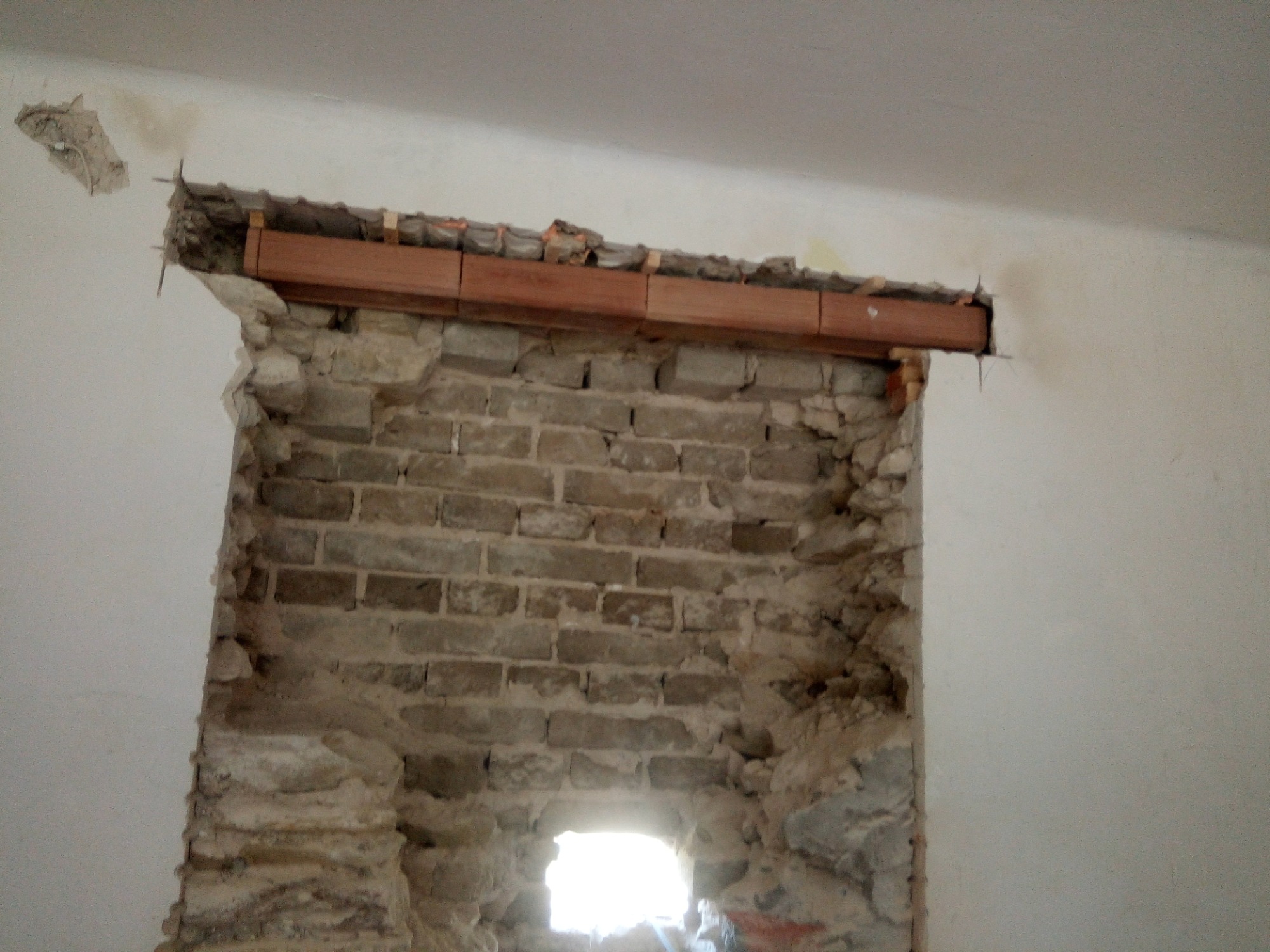 Otwór okienny w trakcie przygotowania: widoczne cegły, drewniana belka podtrzymująca nadproże i fragment oderwanego tynku na ścianie.