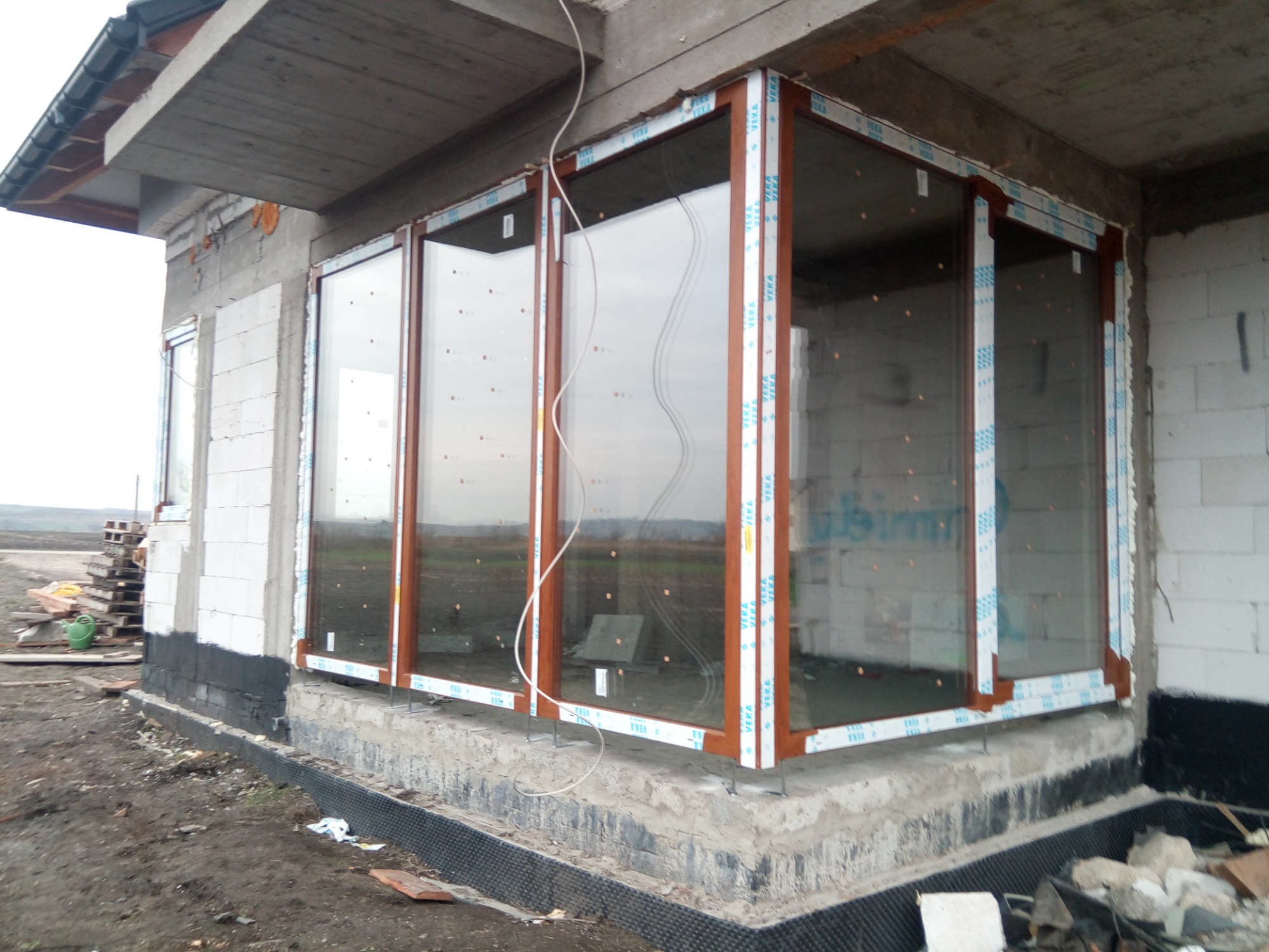 Montaż dużych okien panoramicznych z brązowymi ramami w surowym stanie budynku, widoczne zabezpieczenia na ramach i otaczający plac budowy.