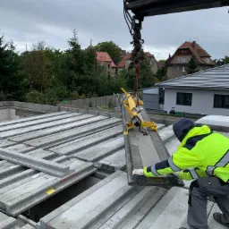 Montaż prefabrykowanych stropów Teriva za pomocą dźwigu i chwytaka, pracownik w odblaskowej kurtce, widok na domy w tle.